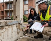 This image shows a construction safety inspector inspecting a deteriorating concrete wall at a residential HOA or condo complex. The worker is seen noting measurements and observations on a clipboard while another official records details nearby.